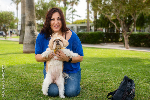 Young woman kneels on green grass in a sunny park. She smiles while holding a fluffy Shih Tzu. The dog looks toward the camera with bright eyes. Black bag rests on the grass nearby