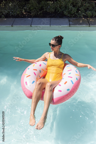Adult woman is floating on pink sprinkled donut float in light-blue pool, sunlight sparkling