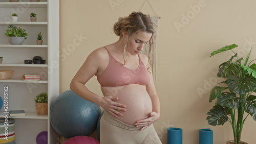Woman cradling pregnant belly with hands  wearing sports bra and leggings in yoga studio  serenity anticipation.