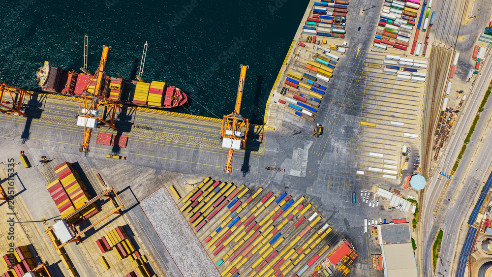 Obraz premium Aerial view of a global logistics hub with a massive container ship being loaded by cranes at a busy port terminal amid rows of colorful shipping containers and quay activity