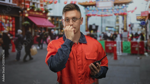 Vászonkép Man in orange coverall holding a walkie, hand to mouth biting nails, looking tense in a crowded street festival; urgent concern