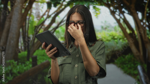 Bild auf Leinwand Woman pinching nose and holding tablet on forest path under trees, wearing glasses and green shirt; disgust