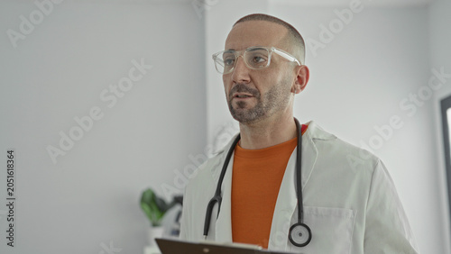 Vászonkép Man doctor with stethoscope and labcoat reading a clipboard while speaking and wearing clear glasses in clinic; calm care