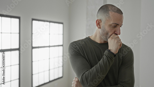 Foto Man with hand on chin, thumb near mouth, standing by a tall window in an empty building room, looking thoughtful and pensive; contemplation