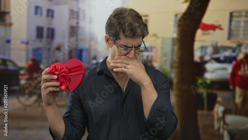 Konstfotografi Man holding red heart gift with hand to mouth on street, examining ribbon and bow with pained expression; heartbreak sadness