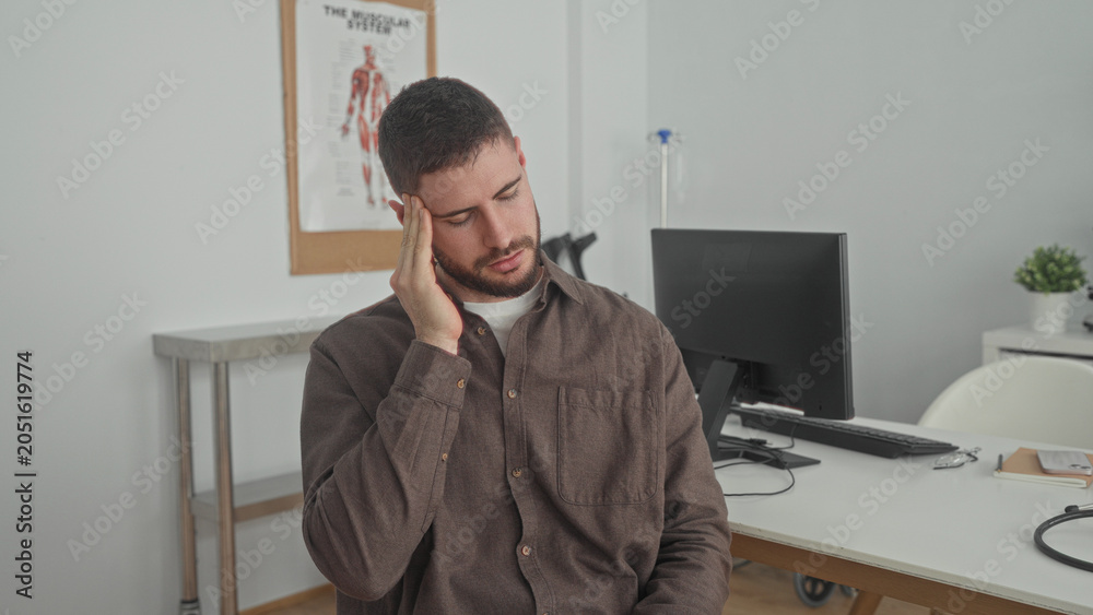 Fototapeta premium Man holding temples with both hands and closed eyes in a clinic building consultation room; pain concern.
