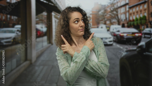 Tableau sur toile Woman pointing both index fingers with crossed arms on a street storefront, shrugging palms and raising eyebrows; confusion doubt