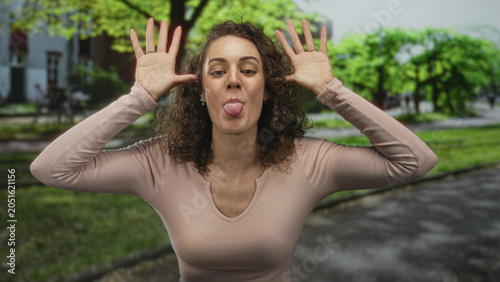 Canvas Print Young hispanic woman sticks out tongue and lifts hands to ears in park near green trees and paved walkway; playful mischief