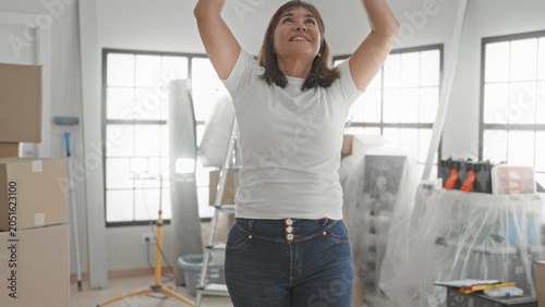 Schilderij op canvas Woman raising arms beside stacked moving boxes and ladder in a bright new building while unpacking and smiling; joy new home beginning