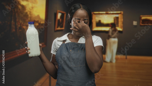 Fotografie Woman wearing apron holds milk bottle and pinches nose, eyes closed, in an art gallery building with framed paintings; disgust reaction