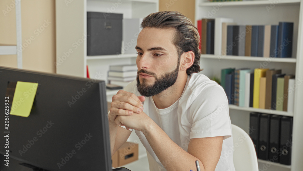 Fototapeta premium Man young hispanic at computer with hands covering face and rubbing jaw while seated at desk in building; stress concentration.