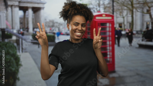 Tableau sur toile Woman smiling and holding up two peace signs with hands on a street by a red phone box; joy celebration