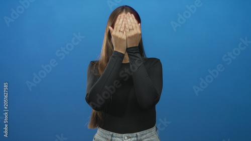 Canvas Print Woman with glasses lifts both hands to frame her face and briefly covers her eyes while pouting in a blue studio; playful curiosity
