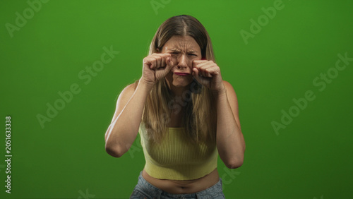 Cuadro en lienzo Woman in yellow crop top and jeans rubs eyes and raises hands near face, showing teary frown in a green screen studio; sadness vulnerability