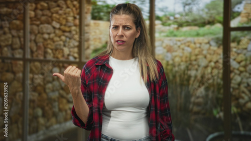 Valokuva Woman pointing thumb over shoulder inside building, wearing red plaid shirt and white tee; annoyance refusal