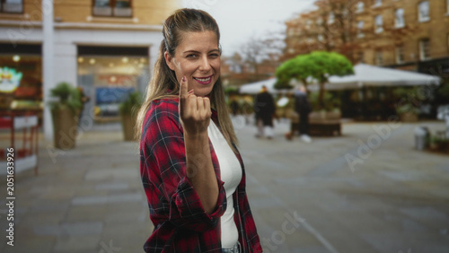 Cuadro en lienzo Woman beckons with finger on street by outdoor cafe and planters, wearing red plaid shirt and white top, smiling at camera; playful invitation
