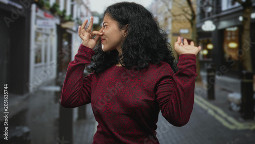 Obraz na plátně Young arab woman pinching her nose with raised hand on crowded city street, wearing red sweater and wincing at a strong smell; disgust avoidance