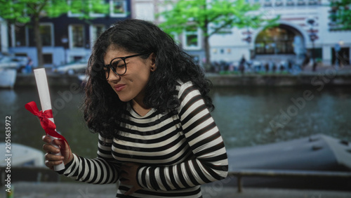 Obraz na plátně Young brunette woman holding diploma with red ribbon and glasses clutching stomach on street by canal, wincing in pain; graduation setback distress