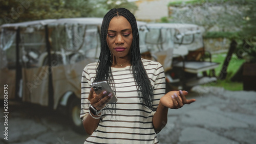 Foto Black woman holding smartphone and frowning while checking phone on street, wearing striped top and smartwatch; frustration solitude