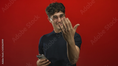Принти на полотні Man holding a smartphone and glancing down in a bright red studio while wearing glasses and a black shirt; curiosity engagement