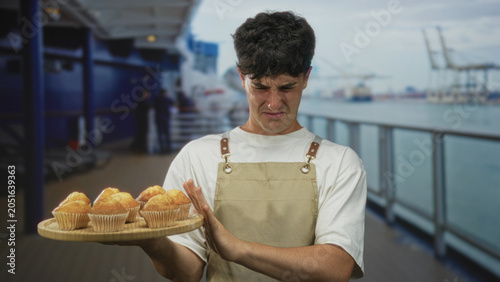 Man wearing apron holding a tray of muffins on a boat deck pushes hand away and grimaces at food; disgust refusal.