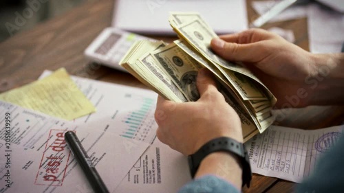 A young businessman's hands tally banknotes beside unpaid bills and scattered documents, the weight of finance pressing down. The scene conveys mounting pressure as he confronts growing obligations