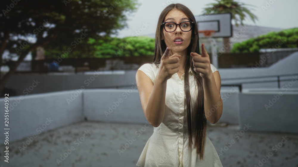 Fototapeta premium Woman with long hair and glasses pointing fingers toward camera by a basketball hoop in a building courtyard; surprise curiosity invitation.