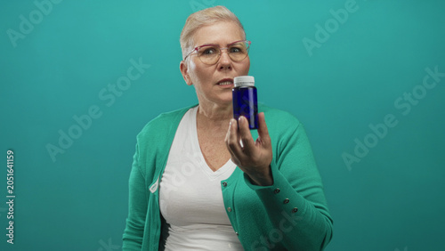 Photography Woman holding cobalt pill bottle with clenched fist, wearing green cardigan and white tee, short hair and glasses on teal backdrop in studio; determination