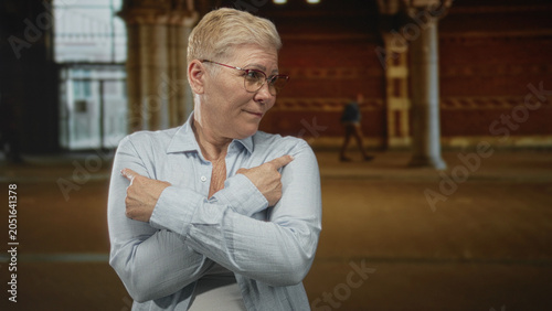Canvas Print Woman wearing glasses with crossed arms pointing fingers outward in a covered building arcade near stone columns; quiet confidence