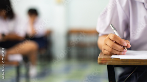 Close-up of a student's hand diligently writing on an exam paper during a school test. 
The student is using a professional pen , sitting at a wooden desk in a brightly lit classroom.