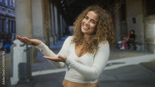 Bild auf Leinwand Young woman with curly hair presenting palms up, smiling, showing navel piercing and white cropped sweater amid street colonnade and stone arches; warm friendly joy