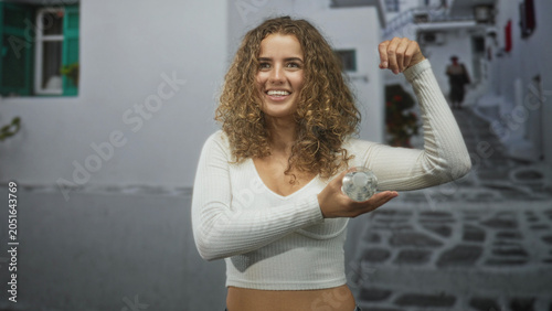 Slika na platnu Woman holding a water bottle in her hand, flexes her arm and shows midriff while smiling on a cobblestone street with whitewashed walls; hydration health playful