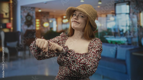 Foto Redhead woman pointing to wrist and tapping with fingers, frowning in a hotel lobby building; impatience dismissal