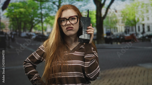 Photos Redhead woman holding vintage cellphone to ear, raising hand on street, wearing striped brown sweater and glasses; retro curiosity confusion