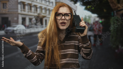 Tableau sur toile Redhead woman wearing glasses holds a retro brick phone to her ear, shrugging with palm up on a city street; frustration disbelief