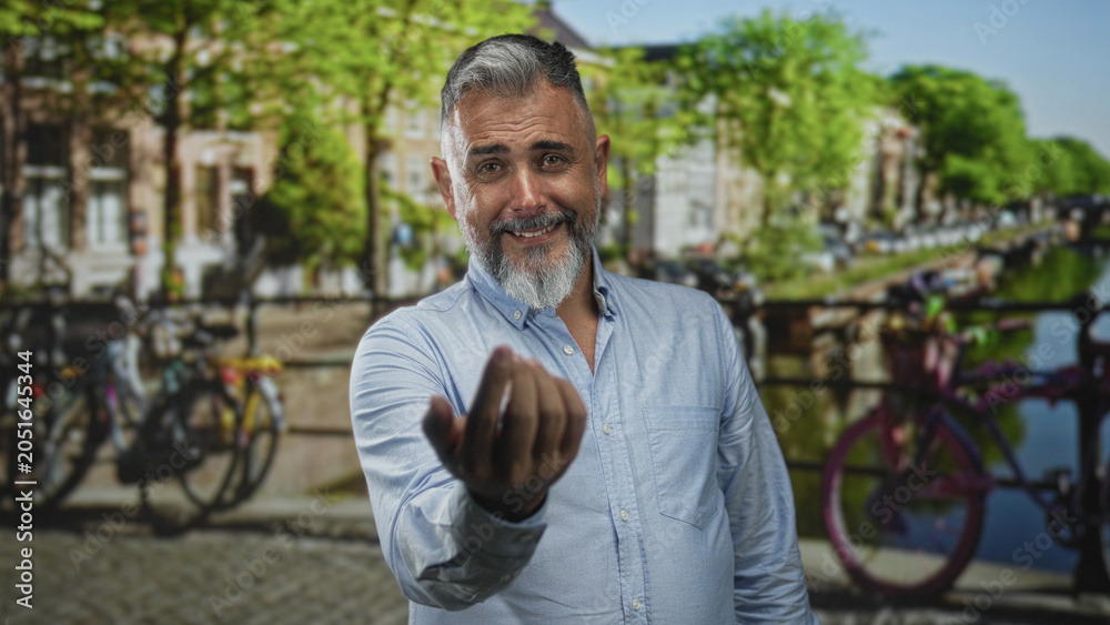 Naklejka premium Man extends hand toward camera beside parked bicycles on amsterdam street while smiling and beckoning; welcoming invitation.