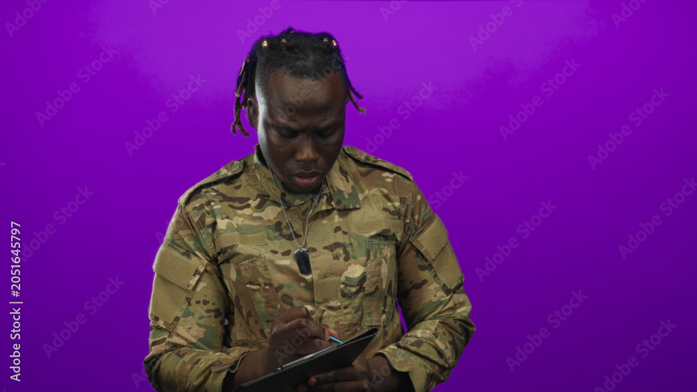 Fototapeta premium Young man in camouflage military uniform with dog tags writes on tablet with stylus and visible hands in purple studio; determination.