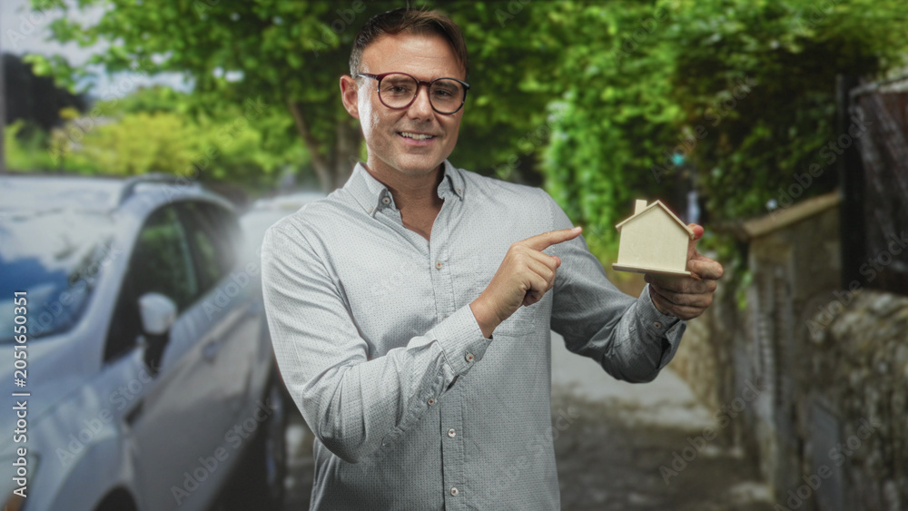 Fototapeta premium Man pointing finger at small wooden house model in residential street, holding model with hand; homeownership pride.