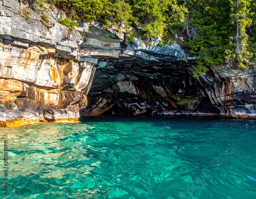 A serene teal-colored body of water in front of a rocky cliffside with lush greenery