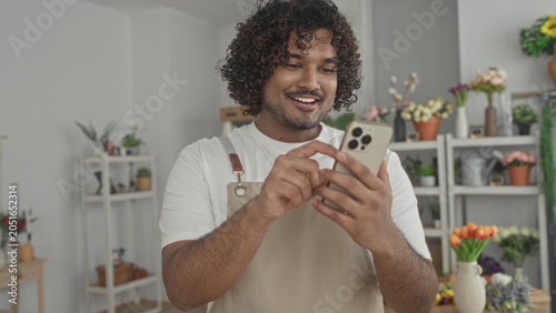 Принти на полотні Man florist tapping smartphone with hands in a flower shop building, wearing apron and smiling at screen; small business care cheerful