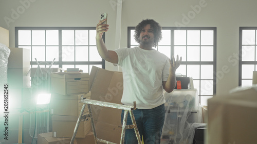 Fotografía Man holds smartphone for selfie with hand on hip amid packed moving boxes and stepladder in building; new home pride