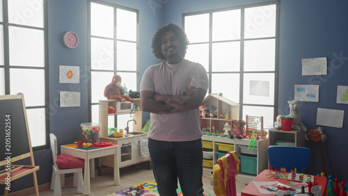 Quadro su tela Man arms crossed standing amid toys and cubbies in a kindergarten building, young teacher smiling at camera; nurturing calm learning