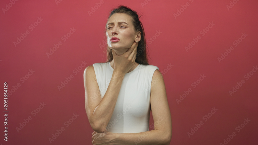 Obraz premium Woman clutching throat and holding neck with one hand in red studio wearing white sleeveless top and closed eyes; pain recovery.