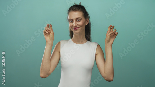 Fotografi Young woman in white sleeveless top making air quotes with both hands and slight smile in teal studio; playful irony