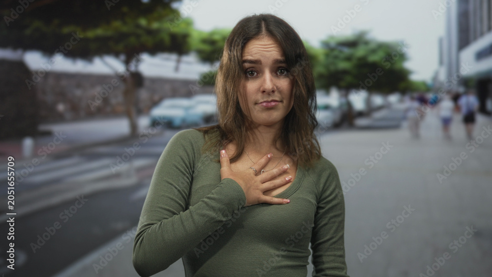 Fototapeta premium Young woman with hand on chest wearing green shirt and necklace on city street pedestrian walkway; regret apology.