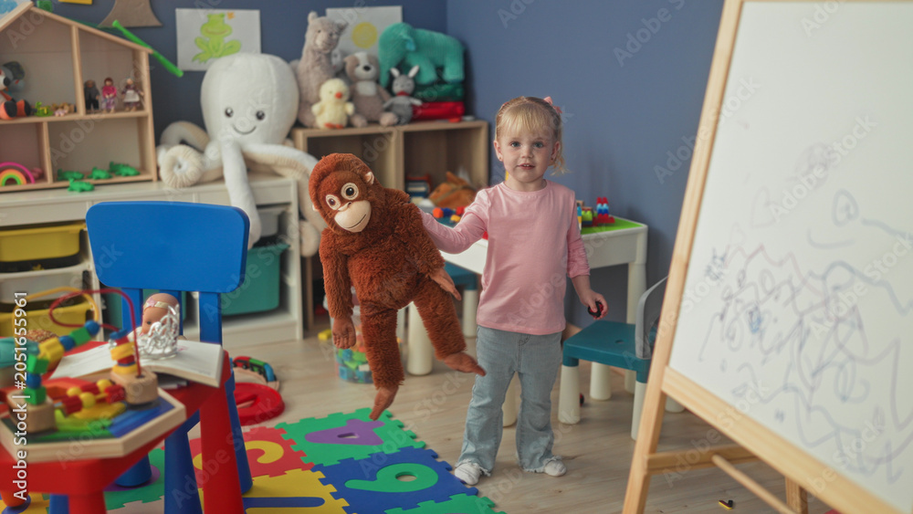 Naklejka premium Little blonde girl hugging a large stuffed monkey with hands in a studio playroom; playful innocence.