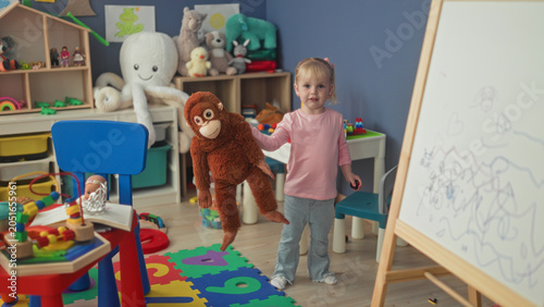 Photography Little blonde girl hugging a large stuffed monkey with hands in a studio playroom; playful innocence