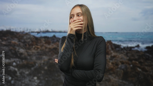 Schilderij op canvas Young hispanic woman in black turtleneck laughing with hand covering mouth and crossed arms on a rocky beach shoreline with ocean waves visible; playful joy
