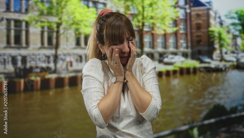 Obraz na plátně Young woman crying with hands covering face in a white blouse on a street in amsterdam; distress urban moment