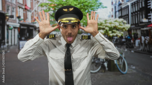 Konstfotografi Man pilot in white uniform and cap pulls hands to ears and sticks out tongue on an urban street with bicycles and storefronts; playful mischief